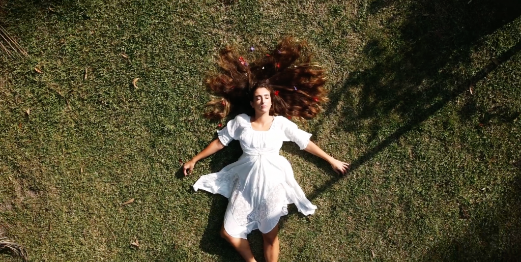 Woman in white dress seen from above lying on grass field her long hair spread around her head and decorated with coloured beads.